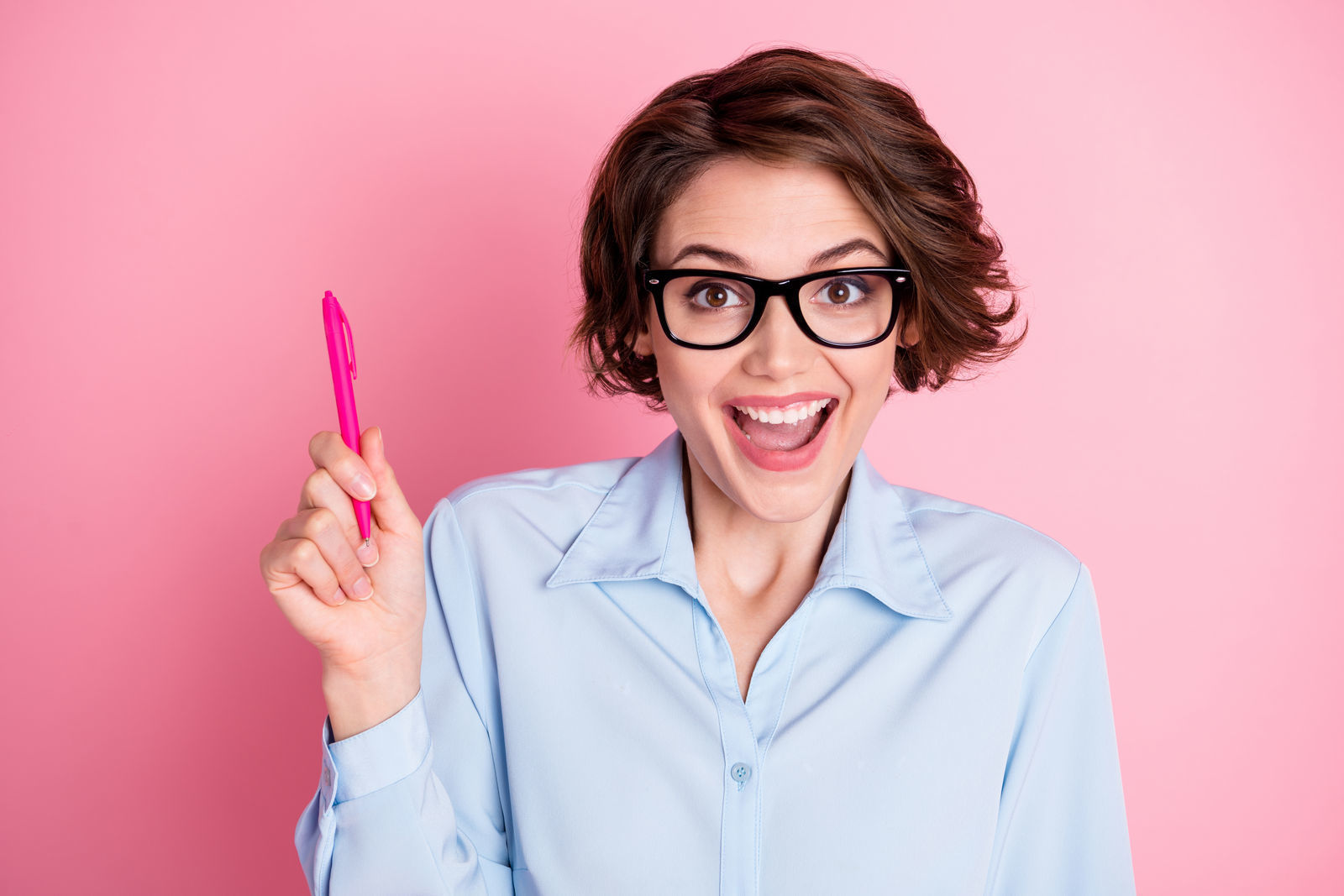 девушка в черных очках с короткой стрижкой в голубой рубашке широко улыбается и держит в руках розовую ручку стоит на фоне розой стены