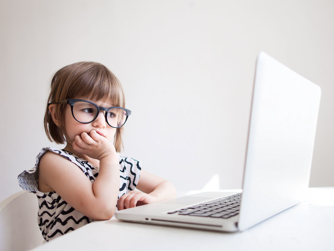маленькая девочка сидит за столом в очках и задумчиво смотрит в ноутбук