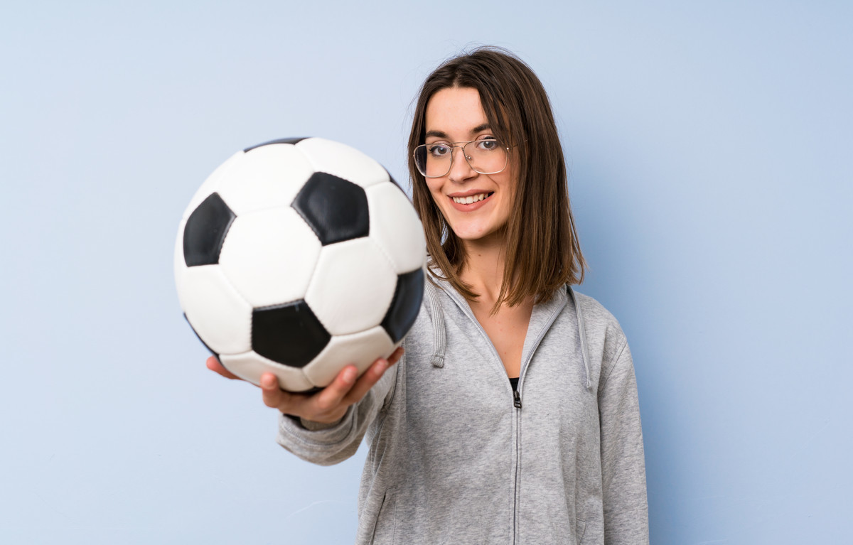 девушка подросток с темными волосами держит на вытянутой руке футбольный мяч