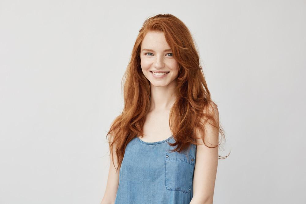 девушка с рыжими волосами стоит на фоне белой стены и улыбается