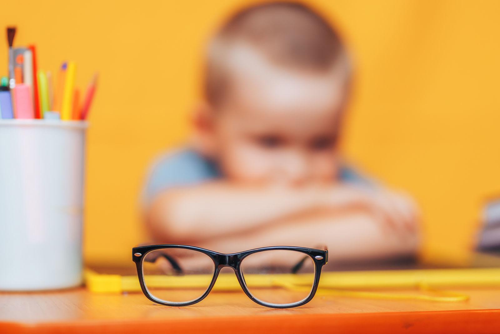 мальчик сидит за столом на фоне оранжевой стены на первом плане очки
