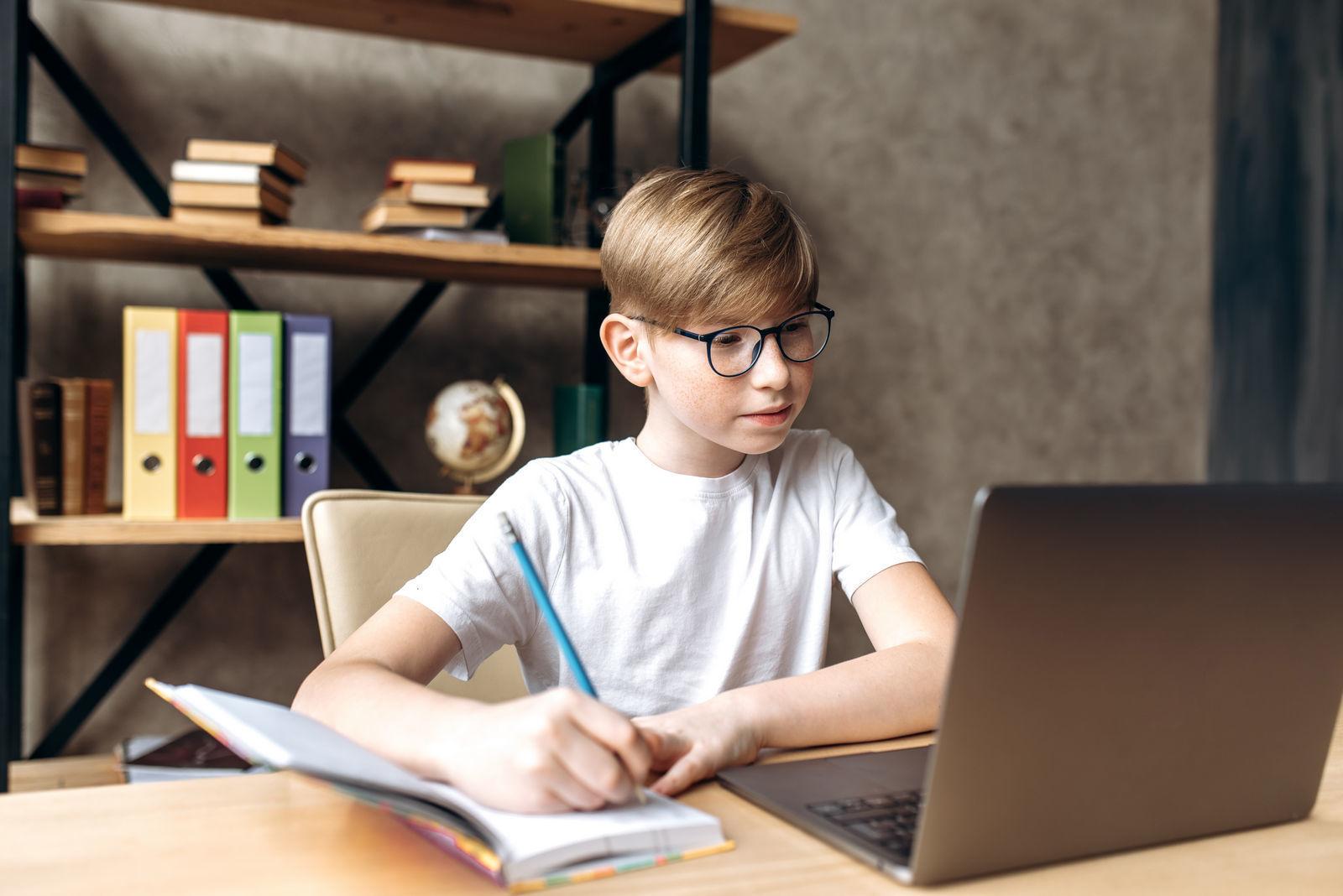 мальчик в очках сидит за учебным столом и делает домашнее задание учится